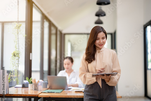 Confident businesswoman holding a clipboard, ready to present in a bright modern office, with colleague working in the background.