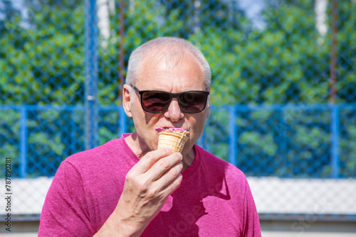 An elderly man in a pink T-shirt eats ice cream on the background of a sports field on a sunny day
