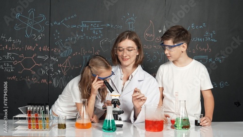 Caucasian boy looking under microscope while teacher giving advice. Professional instructor wearing lab suit looking for diverse student at table with beaker filled with colored solution. Erudition.
