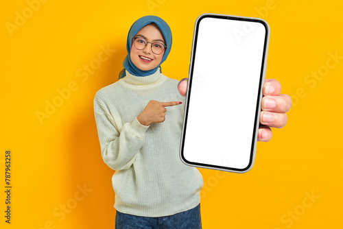 Portrait of cheerful young Asian Muslim woman in white sweater and hijab pointing to blank mobile phone screen isolated on yellow background. Muslim lifestyle concept photo