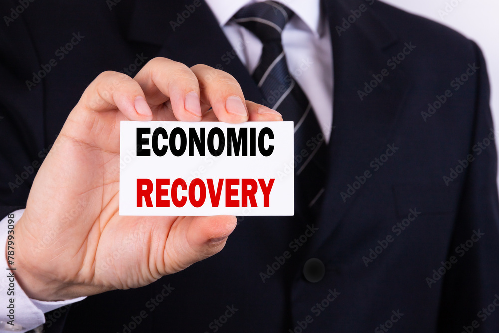 A businessman is holding a tablet with the text Economic Recovery in front of him in his hands. Business concept A businessman is holding a tablet with the text Economic Recovery in front of him in his hands. Business concept