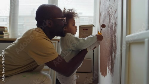 Side footage of caring African American father helping his Biracial child learn to paint walls using paint roller during home redecoration photo