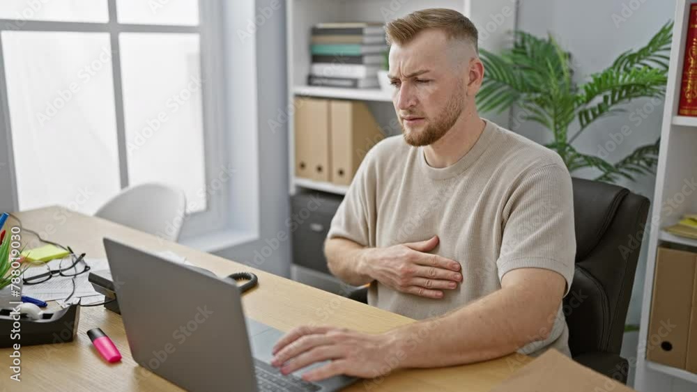 A young bearded man works on a laptop in a modern office, expressing discomfort and chest pain in a health concern concept. A young bearded man works on a laptop in a modern office, expressing discomfort and chest pain in a health concern concept.