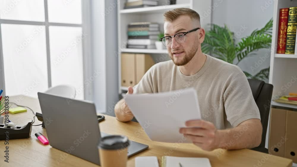 A smiling young bearded man in glasses working on a laptop in a modern office holds papers during a video call. A smiling young bearded man in glasses working on a laptop in a modern office holds papers during a video call.
