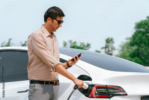 Young man use smartphone to pay for electricity at public EV car charging station green city park. Modern environmental and sustainable urban lifestyle with EV vehicle. Expedient