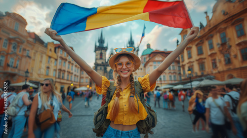 A cheerful young woman dressed in yellow holding a large Romania flag in a historic city square surrounded by people and iconic architecture. photo