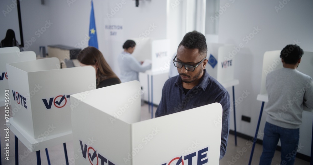 Obraz premium High angle of multiethnic European people voting at polling station during EU elections. Diverse voters fill out ballots in voting booths. Election Day in the European Union. Civic duty and democracy.