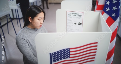 Asian female voter choosing presidential candidate to vote for in voting booth at polling station. US citizen during National Election Day in the United States.