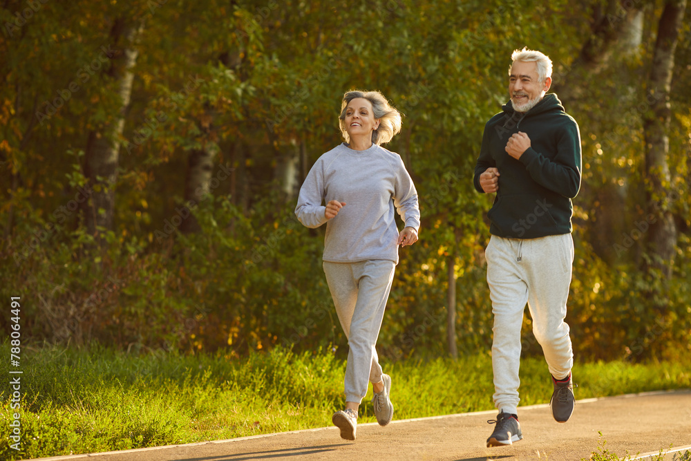 © Studio Romantic - Retirees wife and husband rejoice in active lifestyle. Lovely joyful retirees couple doing sports and jogging together in city park. Senior man and woman in sportswear running on asphalt path.