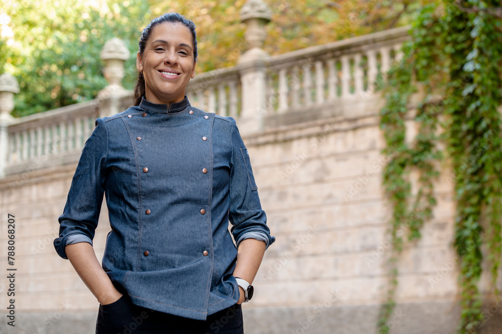 Smiling female chef wears blue coat, poses with hands on pockets outdoors on the street. Restaurant worker, culinary gourmet, pastry chef. Smiling female chef wears blue coat, poses with hands on pockets outdoors on the street. Restaurant worker, culinary gourmet, pastry chef.