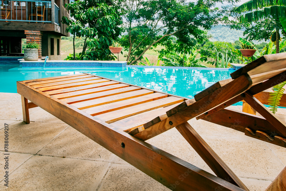 A wooden deck chair near a swimming pool. View of a Deck chair near a beautiful pool surrounded by nature A wooden deck chair near a swimming pool. View of a Deck chair near a beautiful pool surrounded by nature