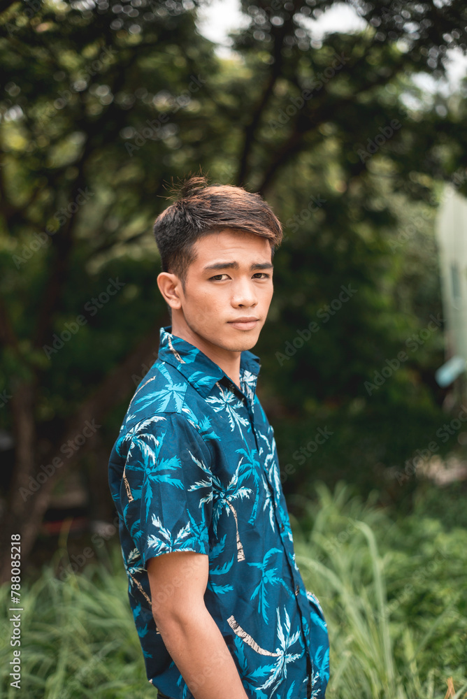 Young man in a vibrant blue Hawaiian shirt standing amid lush green trees Young man in a vibrant blue Hawaiian shirt standing amid lush green trees