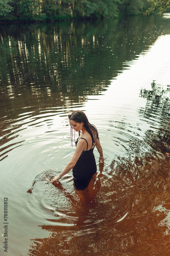 Fototapeta premium Calm woman in wet dress standing in lake and touching water in summer forest