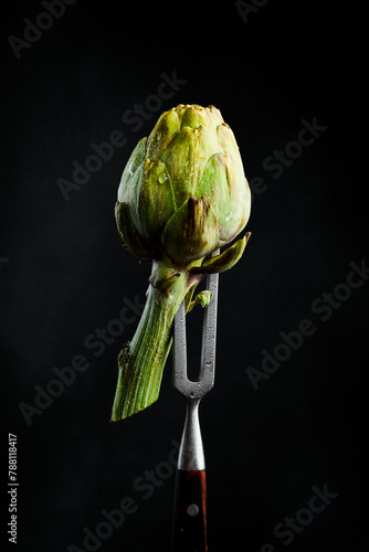 Ripe organic artichoke on a metal fork. On a black background, vertical photo.