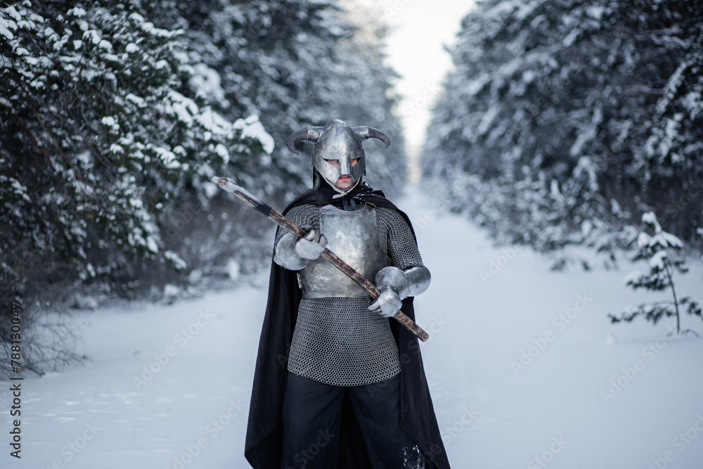 Naklejka premium Portrait of a medieval fantasy warrior in a horned helmet, steel cuirass, chain mail with a two-handed ax in his hands, standing in a fighting position against the backdrop of a winter forest.
