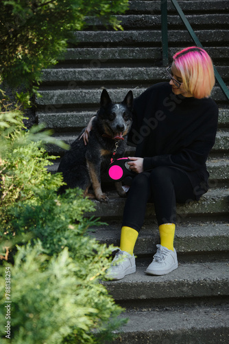 A young attractive woman with pink hair is sitting on the stairs in spring park with her Blue Heeler. Australian cattle dog on a walk with female owner. Family dog outdoor lifestyle concept.