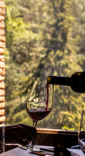 Black wine bottle decanting and pouring red wine into a glass held by a woman's hand backlit by the light of a sunny day coming through the window of a rural town restaurant with the forest behind.