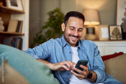 A happy man arranging a meet up with his friends through a text message while sitting on a couch.