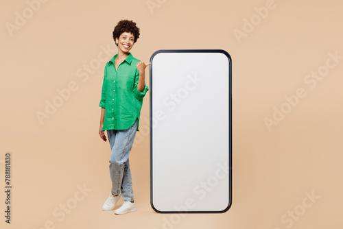 Full body young fun woman of African American ethnicity wear green shirt casual clothes point finger on big huge blank screen mobile cell phone smartphone with area isolated on plain beige background.