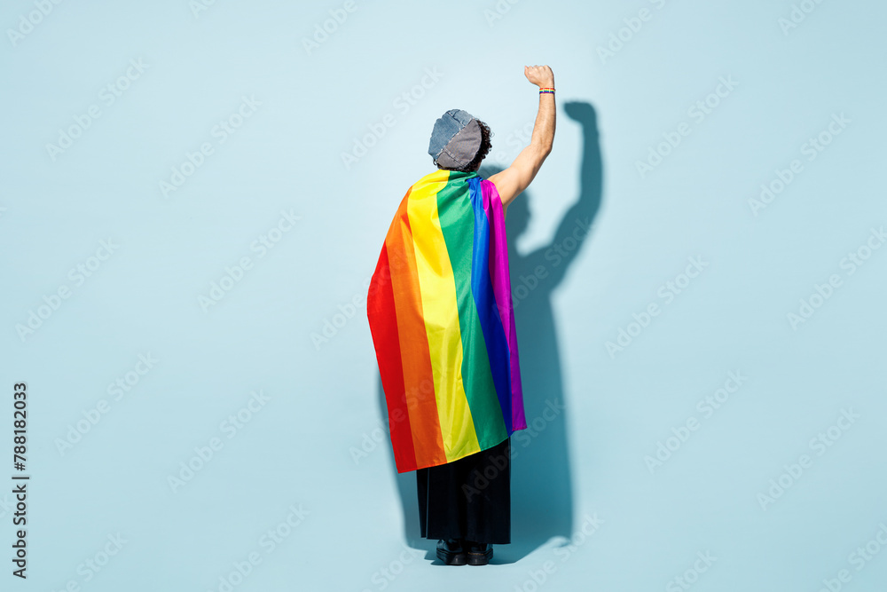 Fototapeta premium Full body back rear view young happy gay man he wrapped in striped rainbow flag clenching fist isolated on plain pastel blue wall background studio portrait. Pride day June month love LGBT concept.