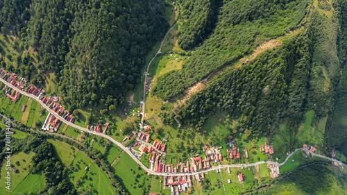Drone panorama above a village located inside a mountainous area, along a river. The river and a road winds near beech forests, and meadows. Countryside landscape, Carpathia, Romania.