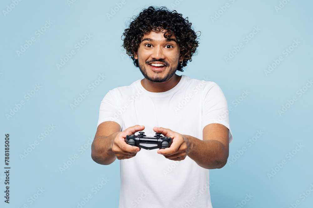Young fun happy Indian man he wear white t-shirt casual clothes hold in hand play pc game with joystick console isolated on plain pastel light blue cyan background studio portrait. Lifestyle concept. Young fun happy Indian man he wear white t-shirt casual clothes hold in hand play pc game with joystick console isolated on plain pastel light blue cyan background studio portrait. Lifestyle concept.
