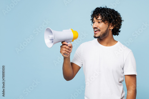 Young happy Indian man wear white t-shirt casual clothes hold in hand megaphone scream announces discounts sale Hurry up isolated on plain pastel light blue cyan background studio. Lifestyle concept.