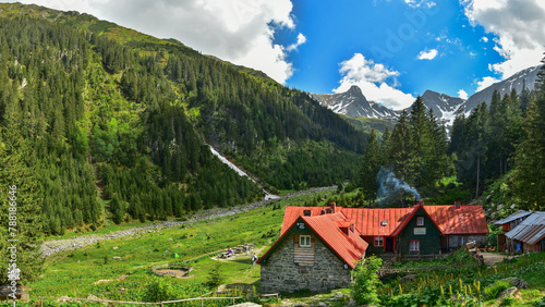 A mountain cabin located inside an alpine meadow, under massive, rocky mountain crests. Lots of hikers are wandering on the trails within the massif. Busy day on the footpaths exiting the forest. 