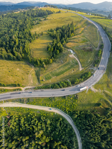 Vertical drone view above a mountain pass, crossing a serene meadow with coniferous forests on it. An asphalt road winds along the mountain peaks. Lots of vehicles are driving on it. Sunset time.