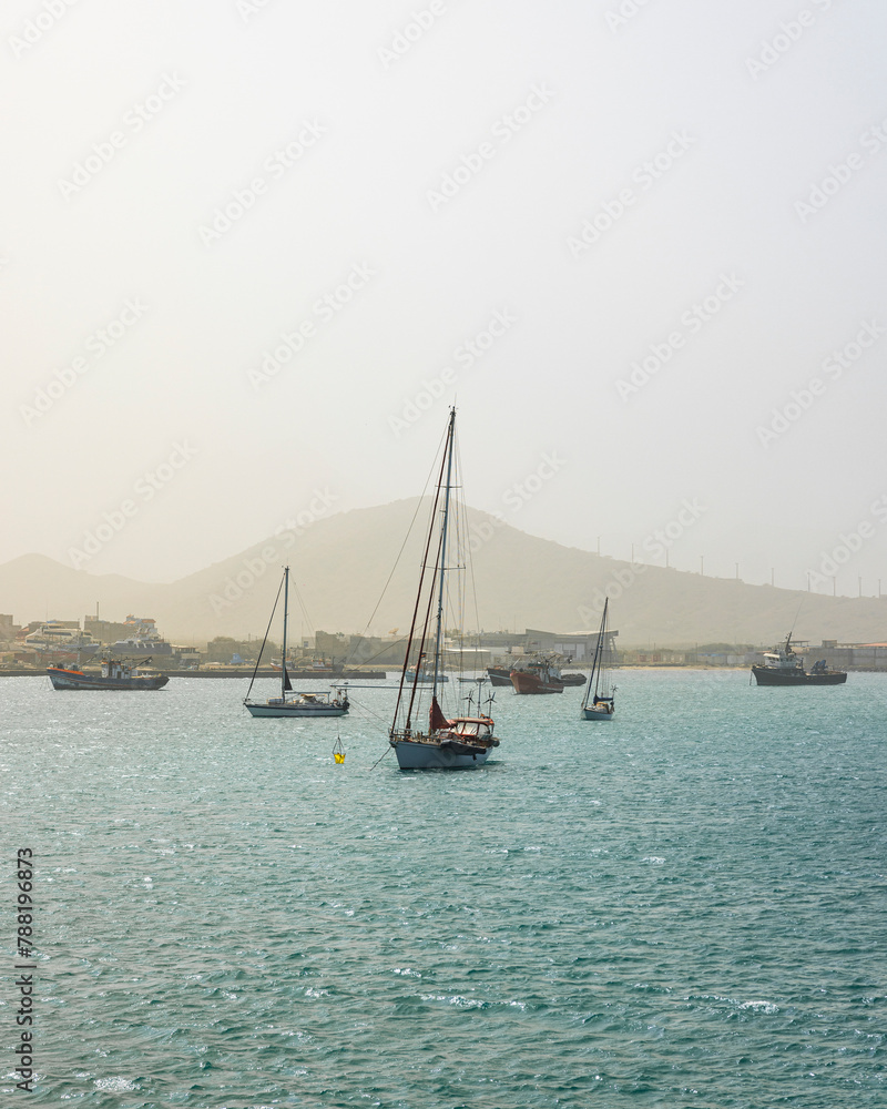 View of a sailing boats in front of the Sao Vicente coastline with mountains during Sahara sandstorm, Cape Verde. View of a sailing boats in front of the Sao Vicente coastline with mountains during Sahara sandstorm, Cape Verde.