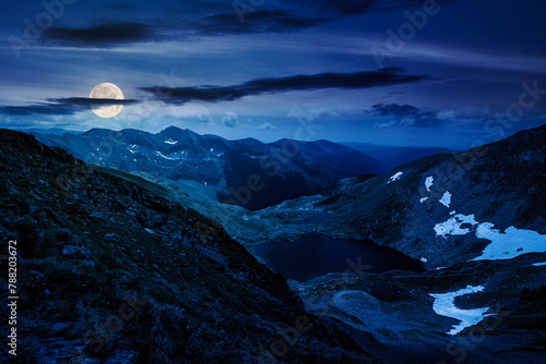 alpine landscape of fagaras mountains at night. capra lake of romania in summer. spots of snow and grass on the rocky hillside in full moon light. stunning travel destination