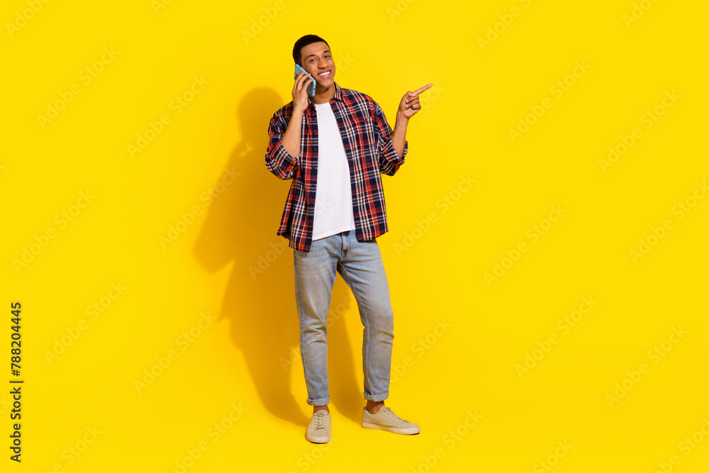 Fototapeta premium Full size photo of attractive man dressed plaid shirt talk on smartphone direting at offer empty space isolated on yellow color background