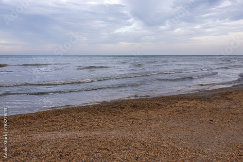 Fototapeta Naklejka Na Ścianę i Meble -  A beach with small shells and the sea. The coast of the Caspian Sea. Cool weather on the coast. A walk along the seashore. The healing sea air. Waves on the surface of the water. Cloudy weather at sea