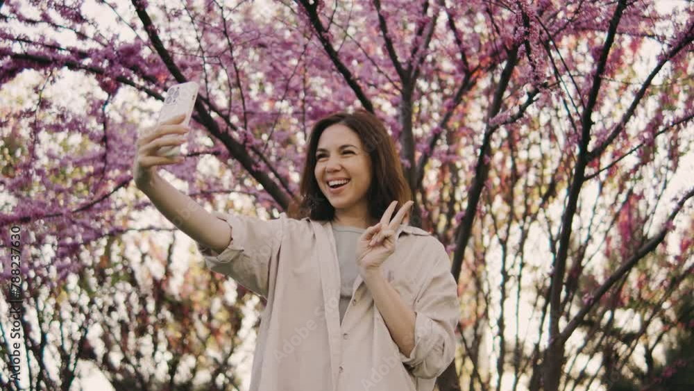 A happy young woman makes a selfie on the background of a tree that blooms with pink flowers. Selfie on a walk, blog, happy moments on camera. Spring flowers of cherry or sakura blossoms on background A happy young woman makes a selfie on the background of a tree that blooms with pink flowers. Selfie on a walk, blog, happy moments on camera. Spring flowers of cherry or sakura blossoms on background
