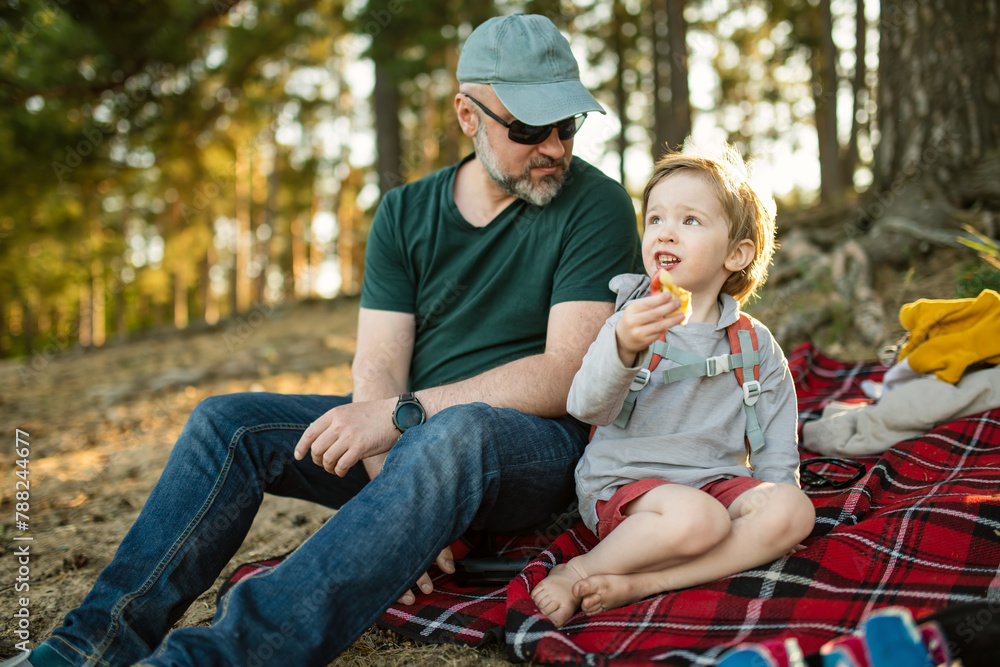 Young father and his little son having a picnic on the lake shore under pine trees. Dad and his boy having good time bonding outdoors. Young father and his little son having a picnic on the lake shore under pine trees. Dad and his boy having good time bonding outdoors.