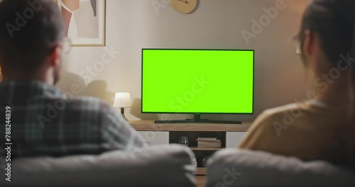 Rear view of married couple sitting at sofa. Young couple sitting in front of TV set with chroma key screen. Wife and husband looking at green mock up screen. Woman and man at living room.