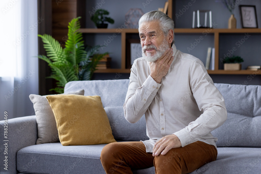 Senior man touching throat while sitting on couch at home Senior man touching throat while sitting on couch at home