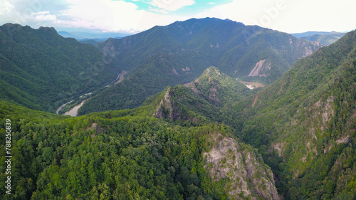 Drone flight above rocky mountain peaks covered with luxuriant and wild beech forests. The valley winds through sharp ravines and gorges. Olt river flows in the background. Carpathia, Romania.