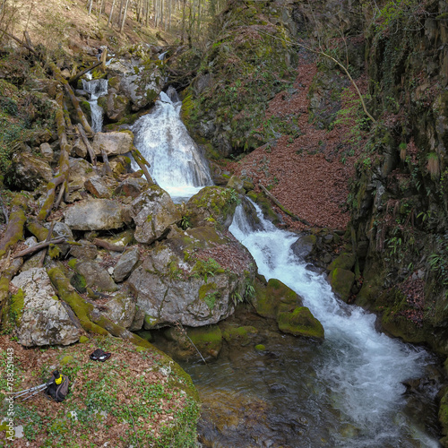 Aerial drone view above a rapid stream flowing through leaves and rocks covered in moss and forming waterfalls. Down on the ground lies photo equipment consisting of a photo camera and a tripod. 