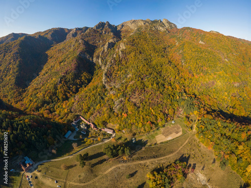 Aerial drone view above a monastery settlement located at the feet of a rocky mountain. The crests are covered with wild beech forests. Autumn season. The woodland is bursting with colour. Carpathia.