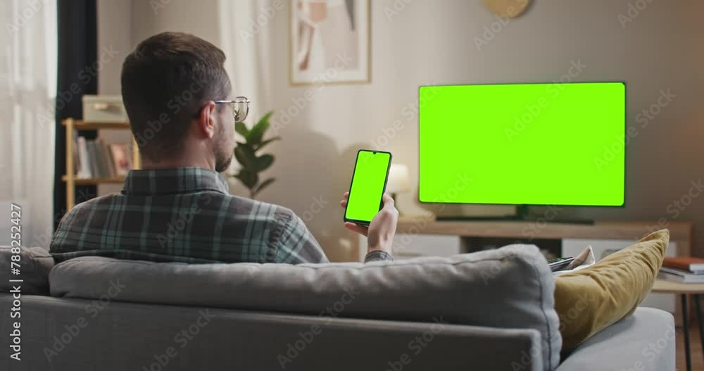 Man holding smartphone with chroma key screen. Male sitting at sofa at background of green mock up TV set. Caucasian man in glasses using green template devices. Man with gadget at modern flat. Man holding smartphone with chroma key screen. Male sitting at sofa at background of green mock up TV set. Caucasian man in glasses using green template devices. Man with gadget at modern flat.