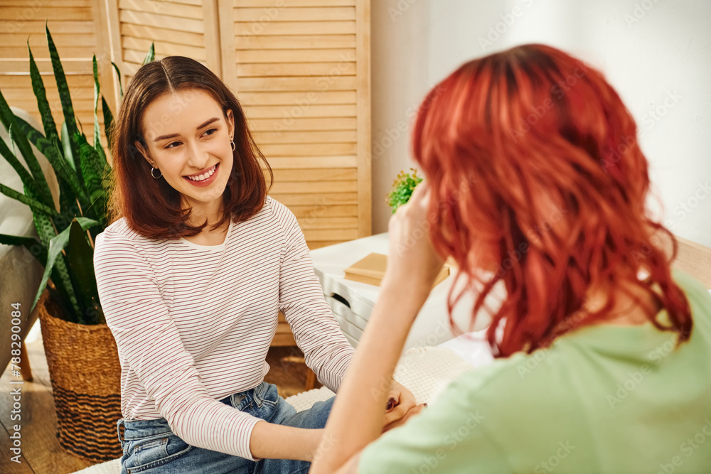 happy young lesbian woman looking at her girlfriend with red hair and smiling in modern bedroom happy young lesbian woman looking at her girlfriend with red hair and smiling in modern bedroom