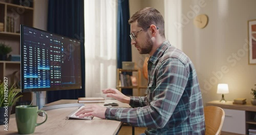 Man sitting in front of computer. Male working at freelance position. Man using computer for cryptocurrency. Male wearing glasses and looking at computer screen. Man working distantly at home. photo