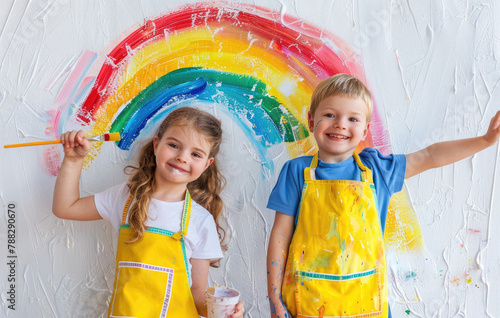 Two children wearing aprons and holding paintbrushes  smiling while painting rainbows on the wall with a white background