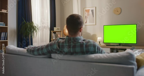 Back view of man sitting at sofa at modern appartment. Male holding remote control. Man looking at chroma key TV set. Male sitting at couch and looking at green moke up TV. Man relaxing at home.