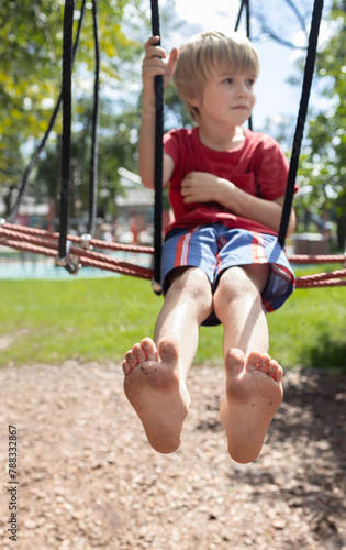 barefoot boy 6-7 years old sits on a sports net in a park on a playground on a summer day. children's playground in the city park, entertainment and recreation for children. Focus on children's feet