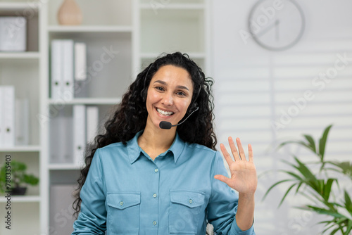 A cheerful woman in a blue shirt and headset waves to the camera, portraying a welcoming online customer service environment in a modern office. © Liubomir