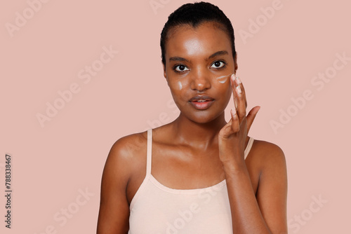 A straight-face African American woman wearing pink top sleeveless is applying moisturizing cream in both face to get ready for work