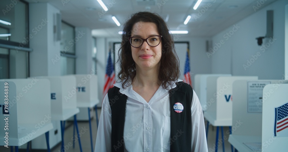 Obraz premium American female voter or polling officer speaks on camera, calls for voting. National Elections Day in the United States. Voting booths in the background at polling station. Civic duty and patriotism.