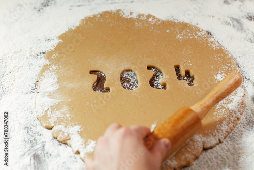 raw dough with 2024 numbers cut from rolled cookie on flour with woman's hand
